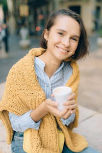 Smiling young woman with a warm drink wrapped in a cozy scarf enjoying a day outdoors.